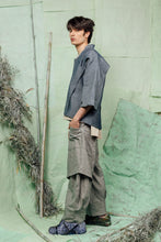 Load image into Gallery viewer, Person wearing a blue shirt and gray pants standing against a textured green wall.